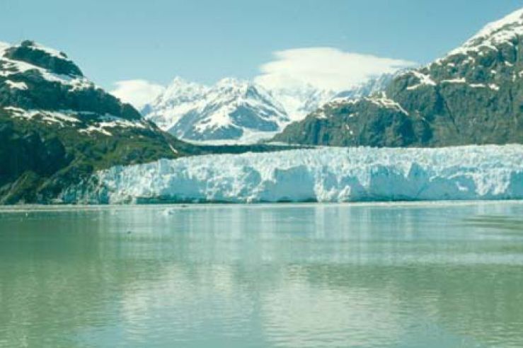 An Alaskan glacier framed by snow-covered mountains, as seen from the opposite side of a glacial Lake.