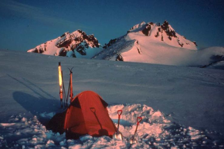 An iconic mountain scene of a small tent pitched amid the snow, looking up slope to rugged snow-covered peaks, drenched in rich evening light.