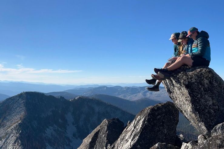 Three people sit on a rocky outcropping on top a mountain ridge